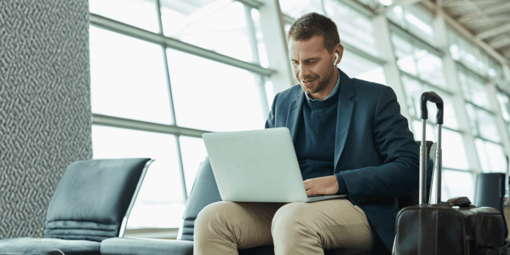 Mann sitzt in einem modernen Flughafenbereich auf einem Stuhl, arbeitet an einem Laptop, neben ihr steht ein schwarzer Rollkoffer.