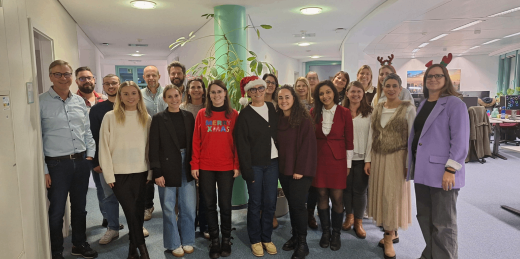 Teamfoto im Büro mit weihnachtlicher Dekoration, Mitarbeitende tragen festliche Kleidung und Accessoires wie Rentiergeweihe und Weihnachtsmützen